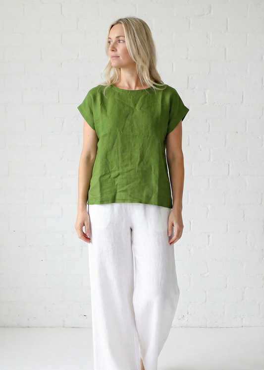 Woman wearing a green top and white pants against a white brick wall.