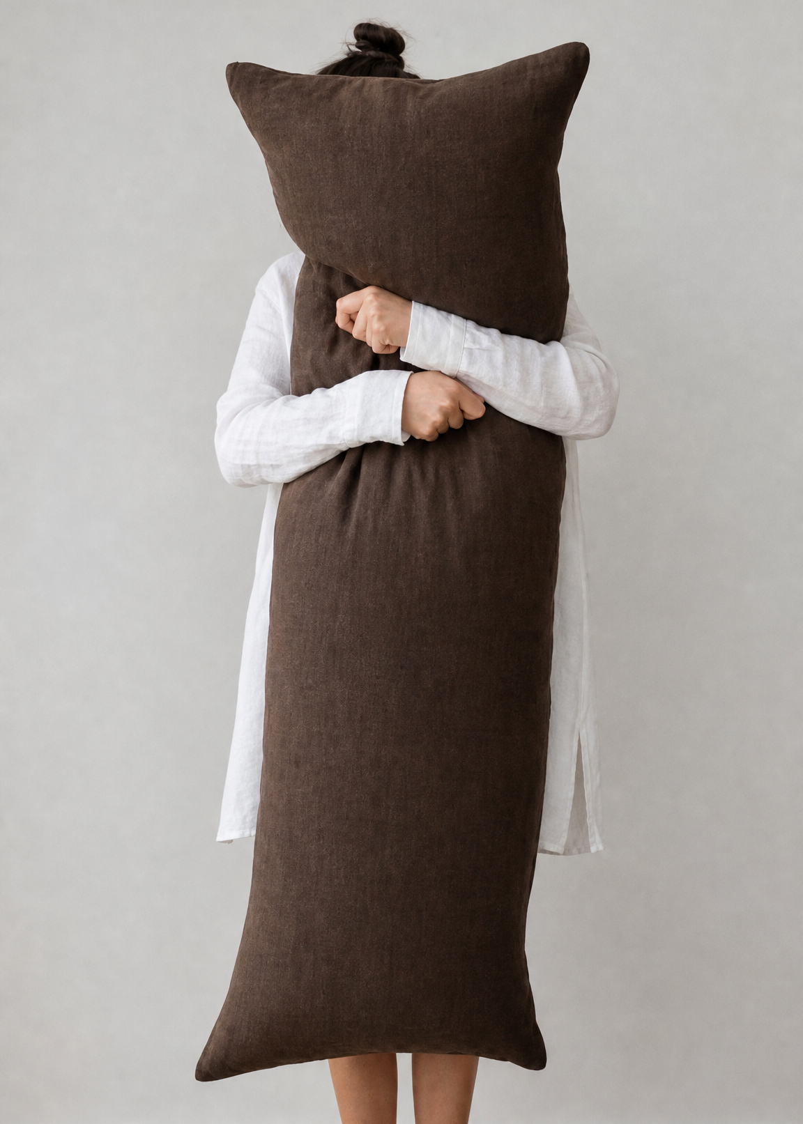 Person wrapped in a long brown towel with a pillow on their head against a plain background