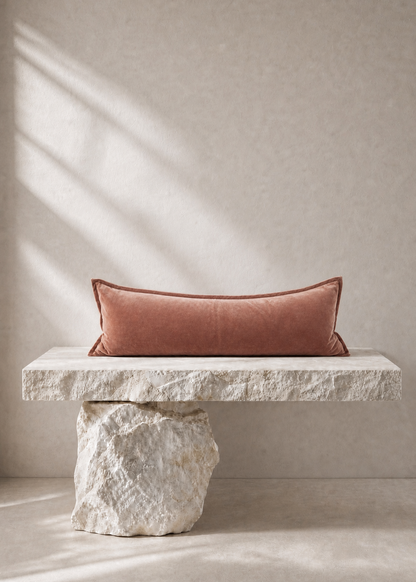 A rectangular dusty rose velvet pillow resting on a rustic stone bench against a textured beige wall with soft window shadow light.