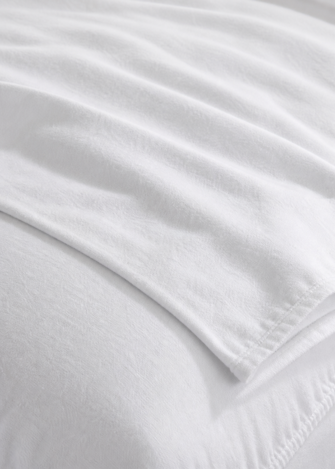 A close-up of a white bed sheet or fitted sheet draped over the corner of a bed, showing the smooth fabric texture and stitched edge.