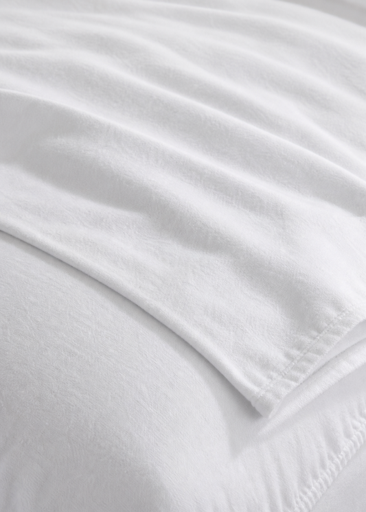 A close-up of a white bed sheet or fitted sheet draped over the corner of a bed, showing the smooth fabric texture and stitched edge.