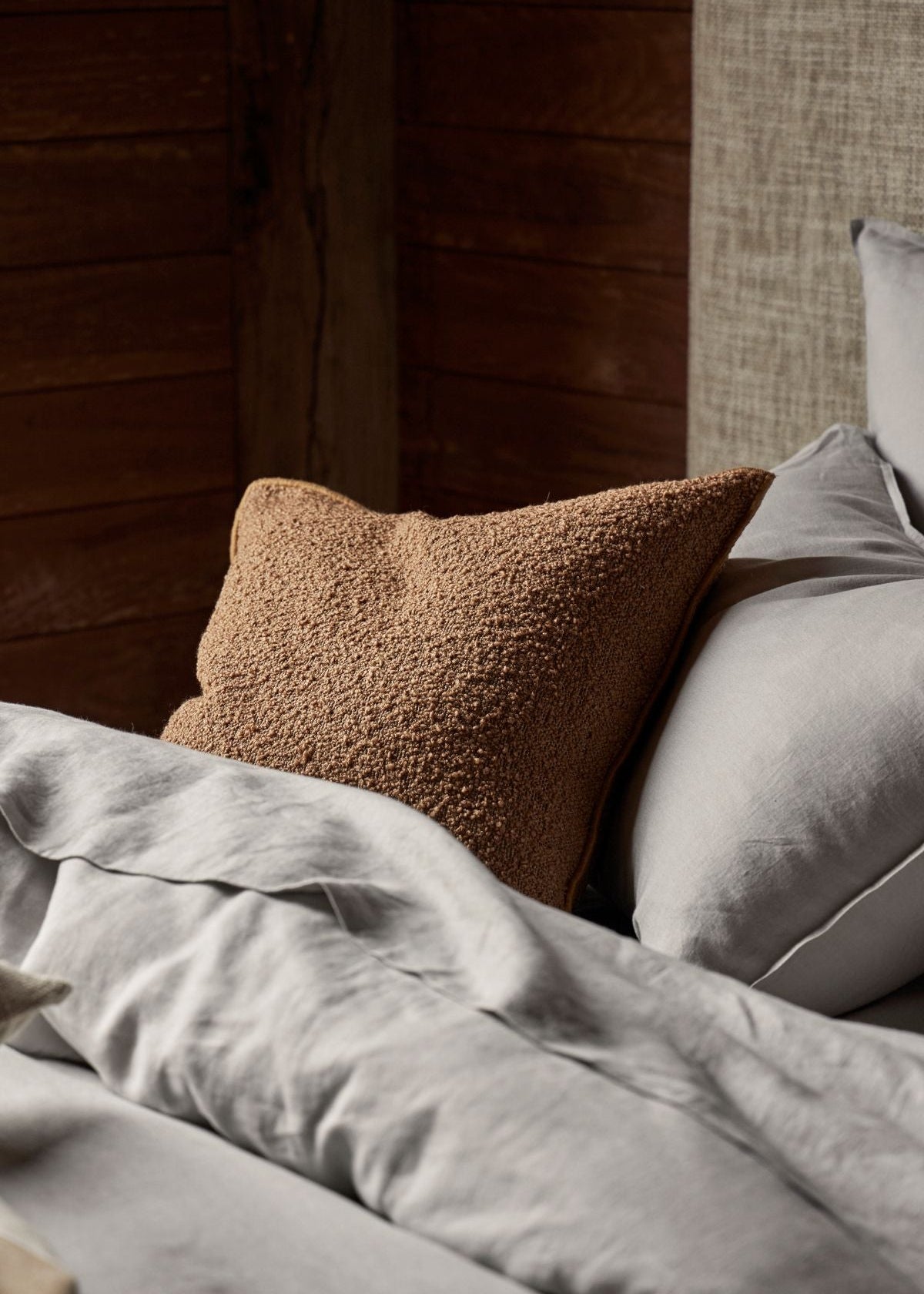 A cozy bedroom scene with a textured brown throw pillow resting on light gray bedding against a dark wood-paneled wall.