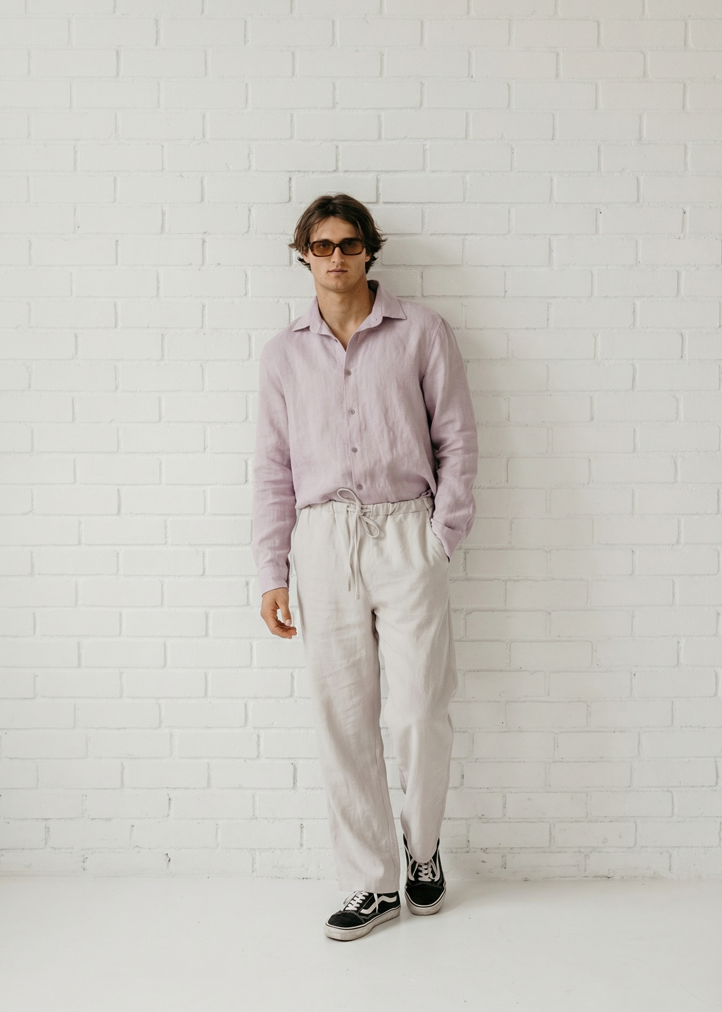 Person wearing a light pink shirt and white pants against a white brick wall.