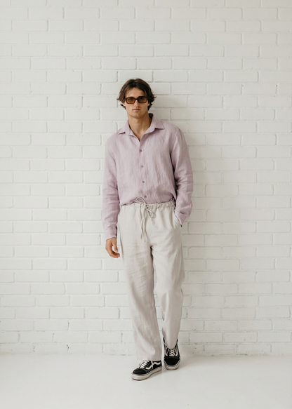 Person wearing a light pink shirt and white pants against a white brick wall.