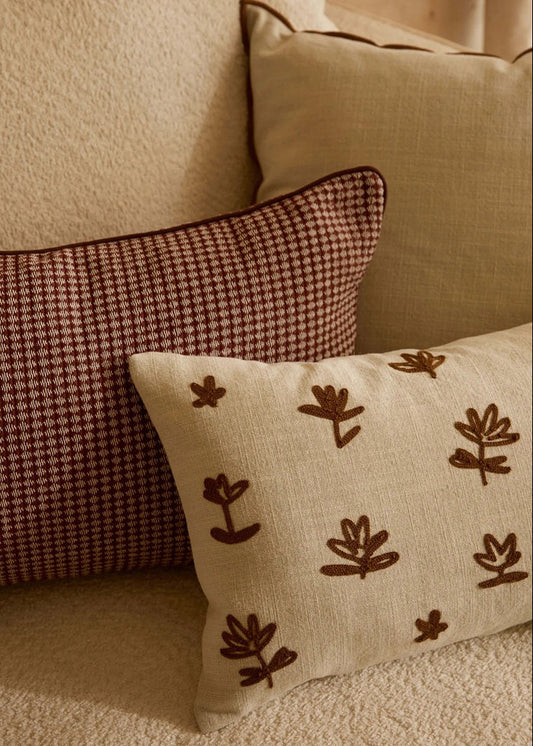 Two decorative throw pillows on a beige sofa: a square pillow with a small-scale brown and beige houndstooth pattern, and a long rectangular pillow in cream linen with brown embroidered botanical motifs.