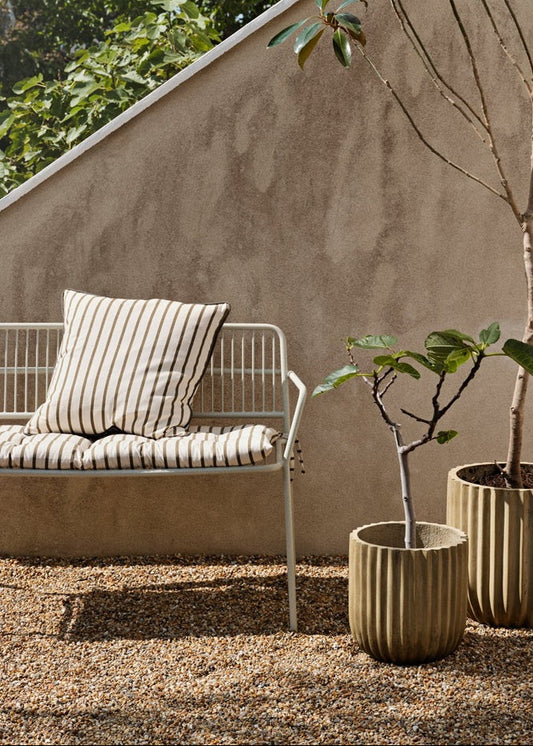 Outdoor setting with a metal bench, striped cushion, and two potted plants against a beige wall.