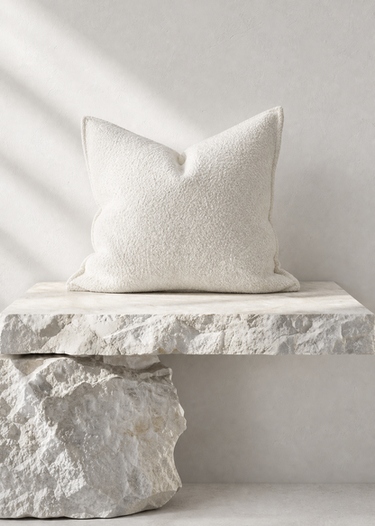 A square off-white textured throw pillow displayed on a rustic white stone bench against a neutral light gray wall with soft diagonal shadows.
