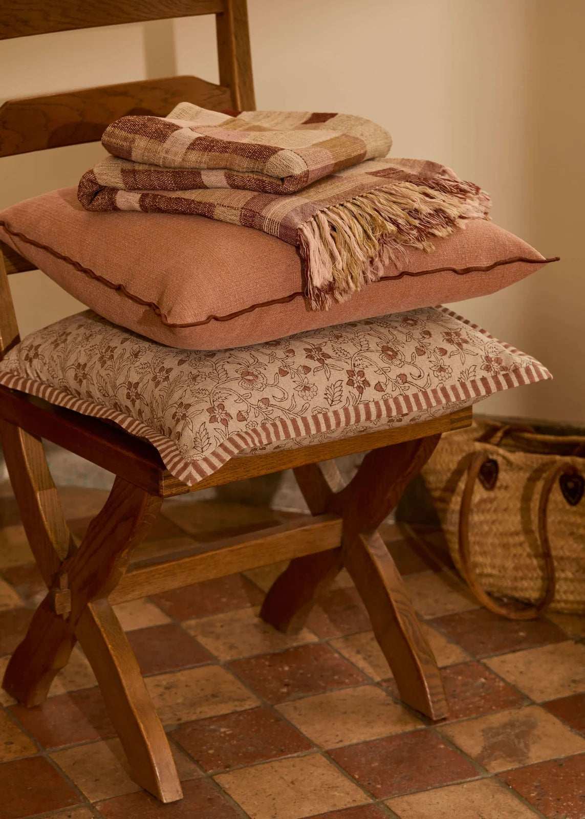 A stack of three decorative cushions and a folded plaid throw blanket arranged on a wooden folding chair in a warm, rustic room with terracotta tile flooring.