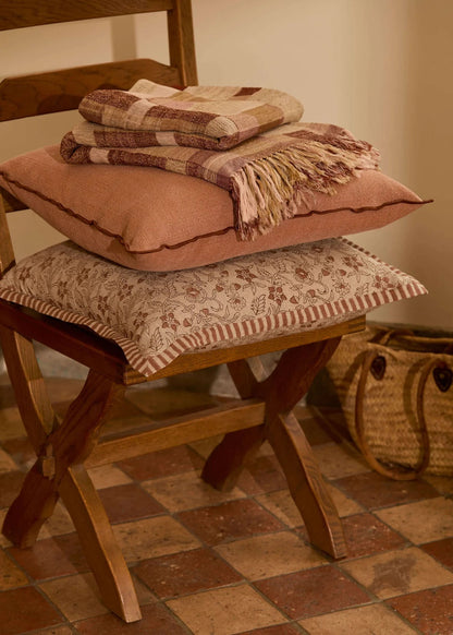A stack of three decorative cushions and a folded plaid throw blanket arranged on a wooden folding chair in a warm, rustic room with terracotta tile flooring.