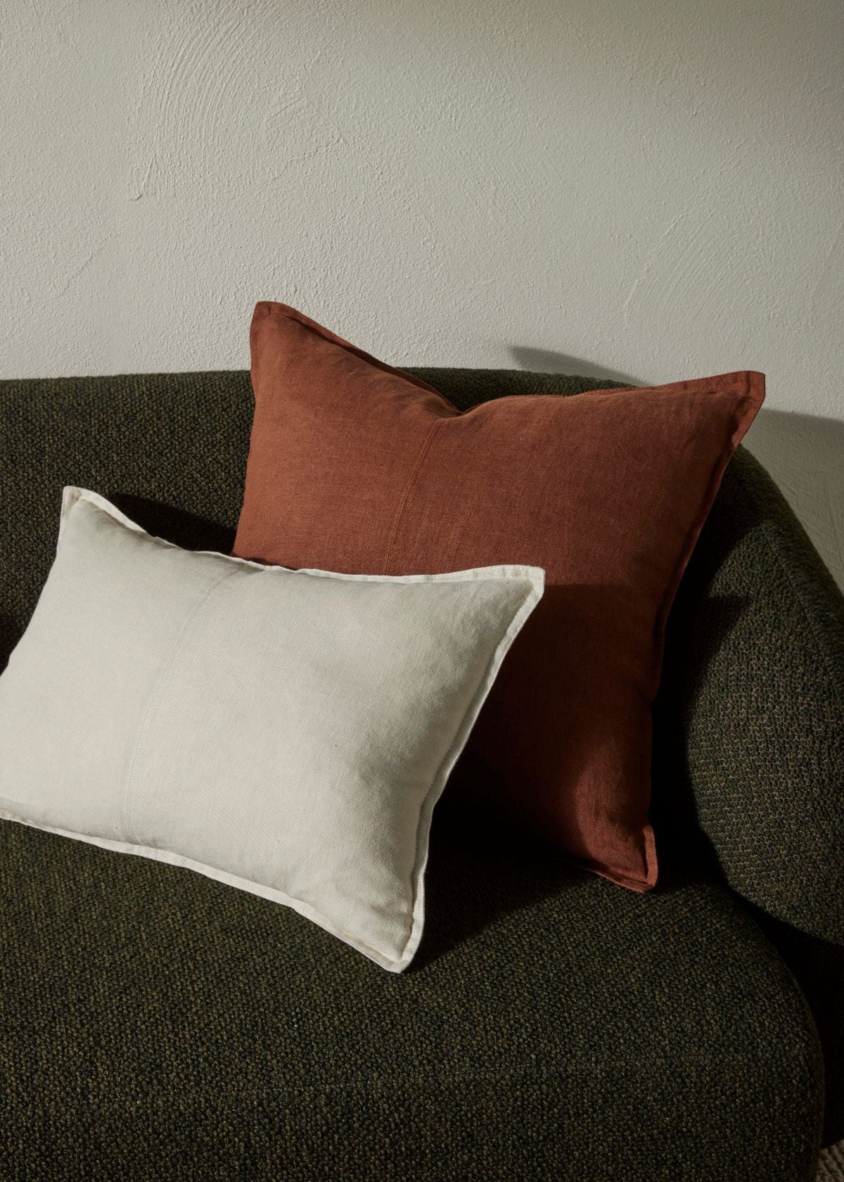 Two decorative throw pillows on a dark textured armchair against a neutral wall. The larger pillow is a warm rust-brown velvet, and the smaller pillow in front is a light cream linen.