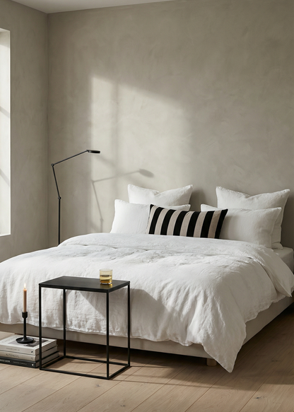 Neatly made bed with white bedding and black and white striped pillow in a room with light-colored walls and wooden floor.