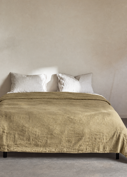 A bed dressed in a textured olive-khaki linen coverlet with two white pillows, set against a warm beige plaster wall and concrete floor.