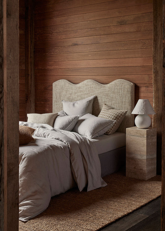 A natural-colored, hand-woven Jute rug with a mirrored stripe pattern, displayed in a bedroom setting with a bed, pillows, and a lamp.