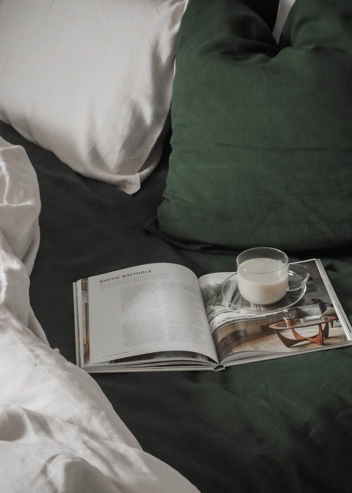A pair of forest green linen pillowcases displayed on a bed with an open book and a cup of coffee on top of the pillowcases.