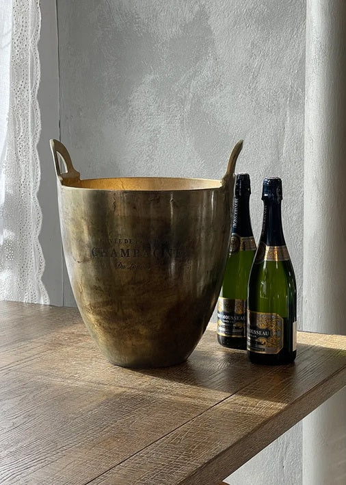 Bronze ice bucket on a wooden surface with champagne bottles in the background.