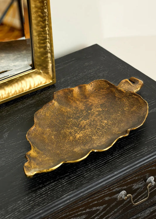 Bronze leaf-shaped tray on a dark wooden surface with a gold-framed mirror in the background.