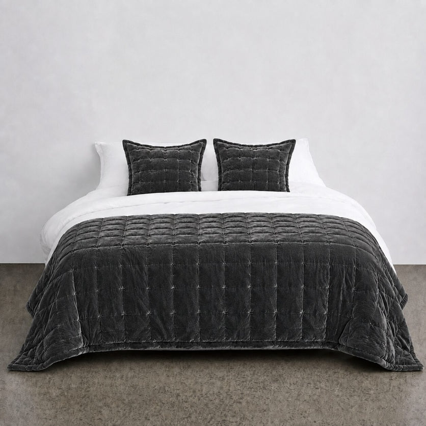 A dark charcoal gray quilted velvet bedding set, including a comforter and two matching pillows, styled on a bed with white sheets against a minimalist gray wall.