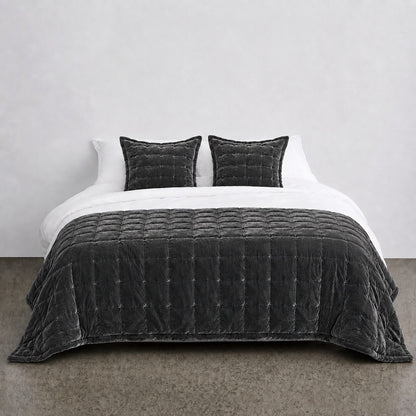 A dark charcoal gray quilted velvet bedding set, including a comforter and two matching pillows, styled on a bed with white sheets against a minimalist gray wall.