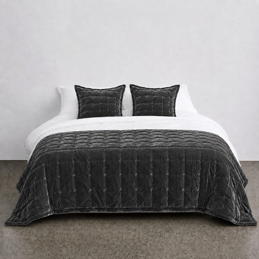 A dark charcoal gray quilted velvet bedding set, including a comforter and two matching pillows, styled on a bed with white sheets against a minimalist gray wall.