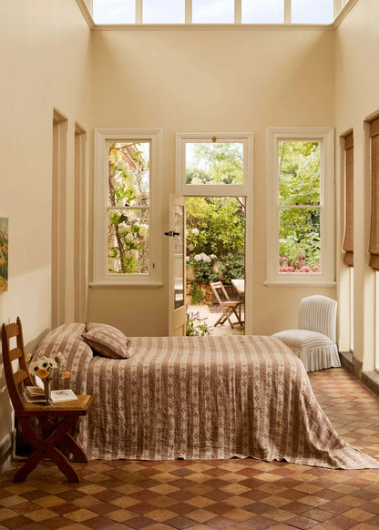 A bedroom interior with a bed dressed in a beige and brown striped quilt, set in a sunlit room with tall windows, a tiled floor, and an outdoor garden visible beyond the glass.
