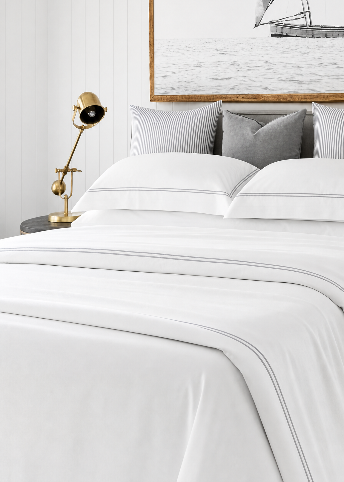 Neatly made bed with white bedding and gray pillows, side table with lamp, and framed artwork on the wall.