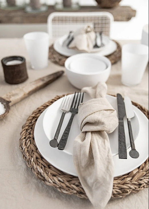 Table setting with woven placemat, white plates, black cutlery, and beige napkin.