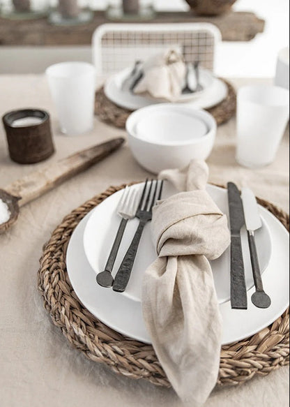 Table setting with woven placemat, white plates, black cutlery, and beige napkin.