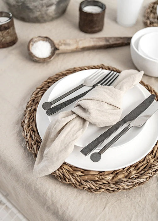 Dining table setting with white plates, black cutlery, and woven placemats.