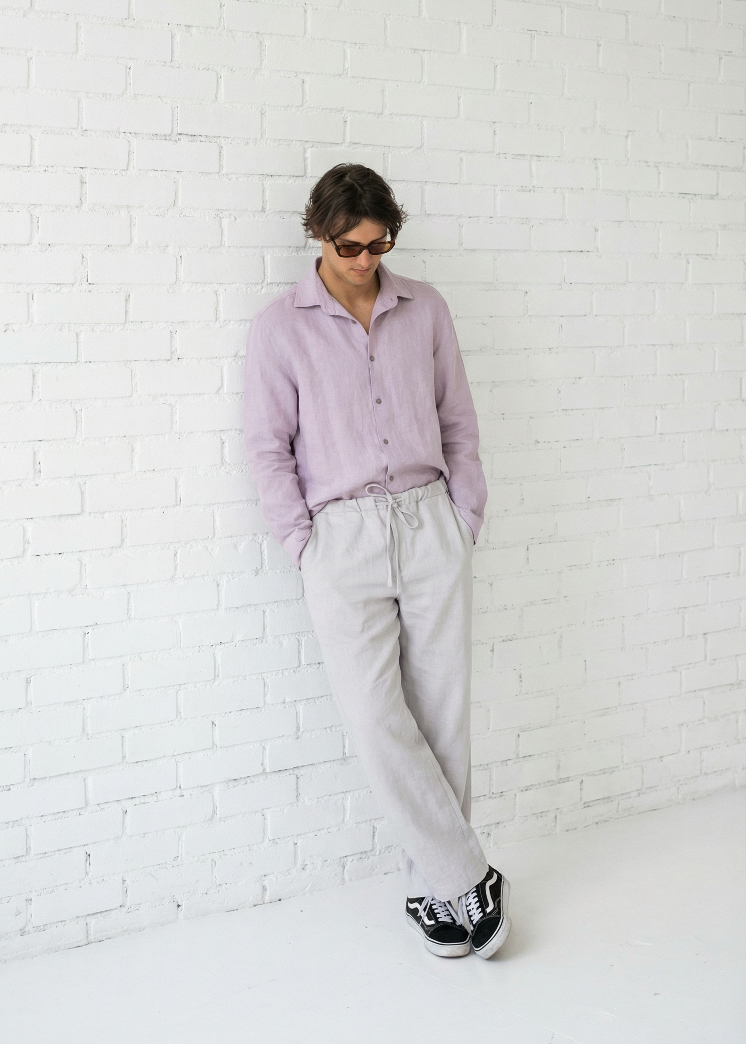 Person wearing a light purple shirt and white pants against a white brick wall.