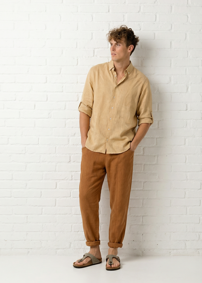 Man wearing a beige shirt and brown pants standing against a white brick wall.