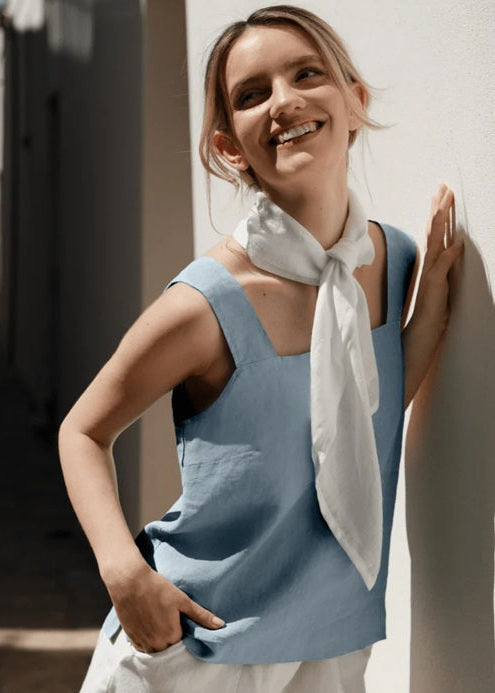 Woman in a blue sleeveless top with a white scarf standing against a white wall with ocean view.