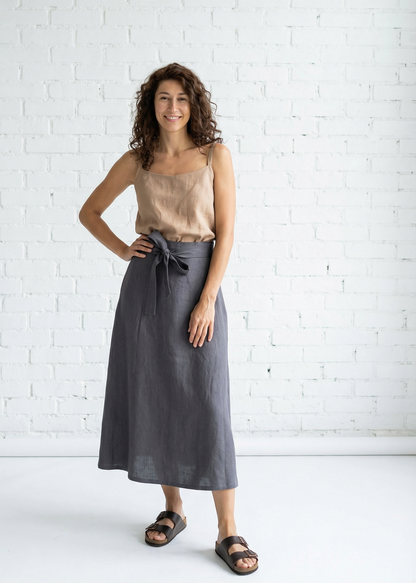 Woman wearing a beige top and gray skirt standing against a white brick wall.