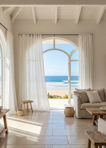 Beachfront living room with large arched window, white curtains, and beige sofa.