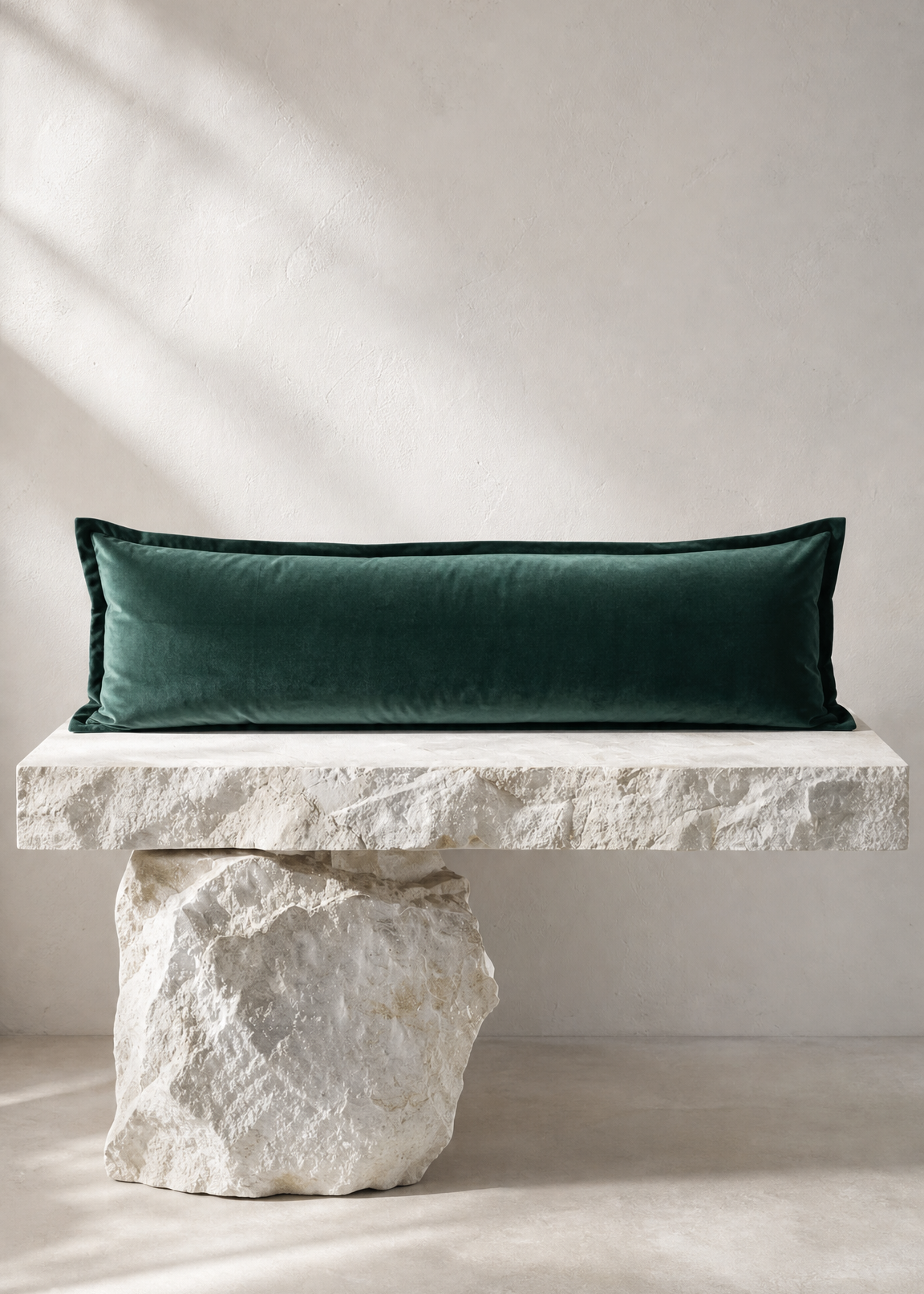 A long rectangular dark green velvet pillow displayed on a white stone bench against a neutral beige wall with soft diagonal shadows.