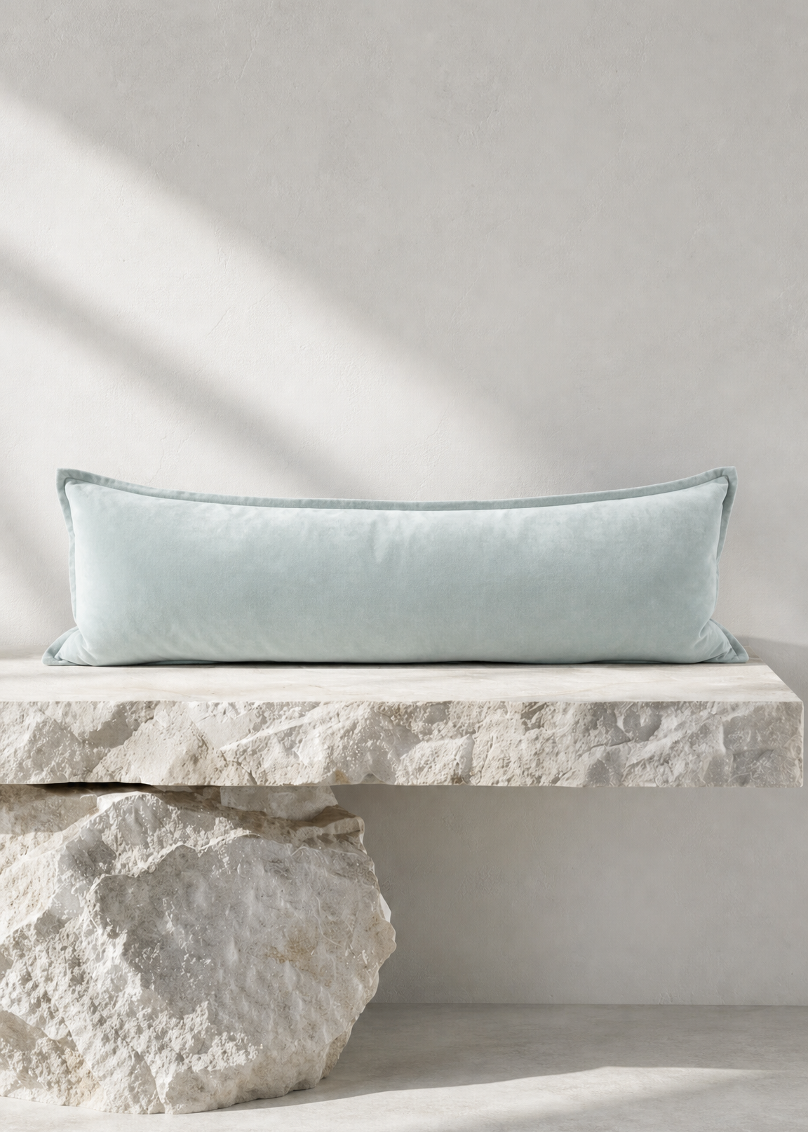 A long rectangular light blue throw pillow resting on a stone bench against a textured light gray wall, with natural shadows cast across the scene.