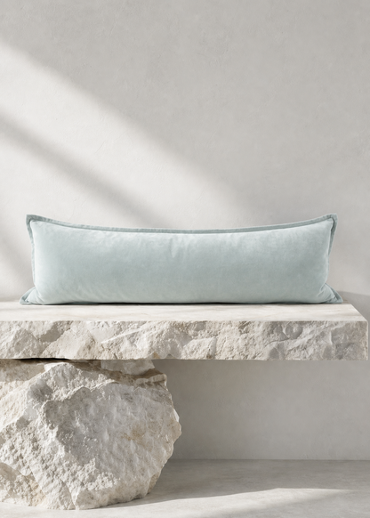 A long rectangular light blue throw pillow resting on a stone bench against a textured light gray wall, with natural shadows cast across the scene.
