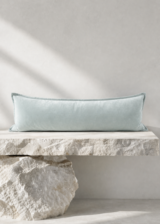 A long rectangular light blue throw pillow resting on a stone bench against a textured light gray wall, with natural shadows cast across the scene.