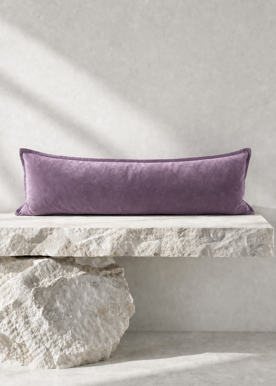 A rectangular lavender velvet lumbar pillow displayed on a rustic white stone bench against a textured light gray wall.