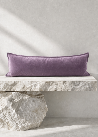 A rectangular lavender velvet lumbar pillow displayed on a rustic white stone bench against a textured light gray wall.