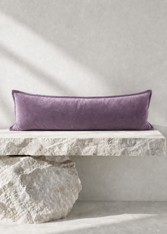 A rectangular lavender velvet lumbar pillow displayed on a rustic white stone bench against a textured light gray wall.