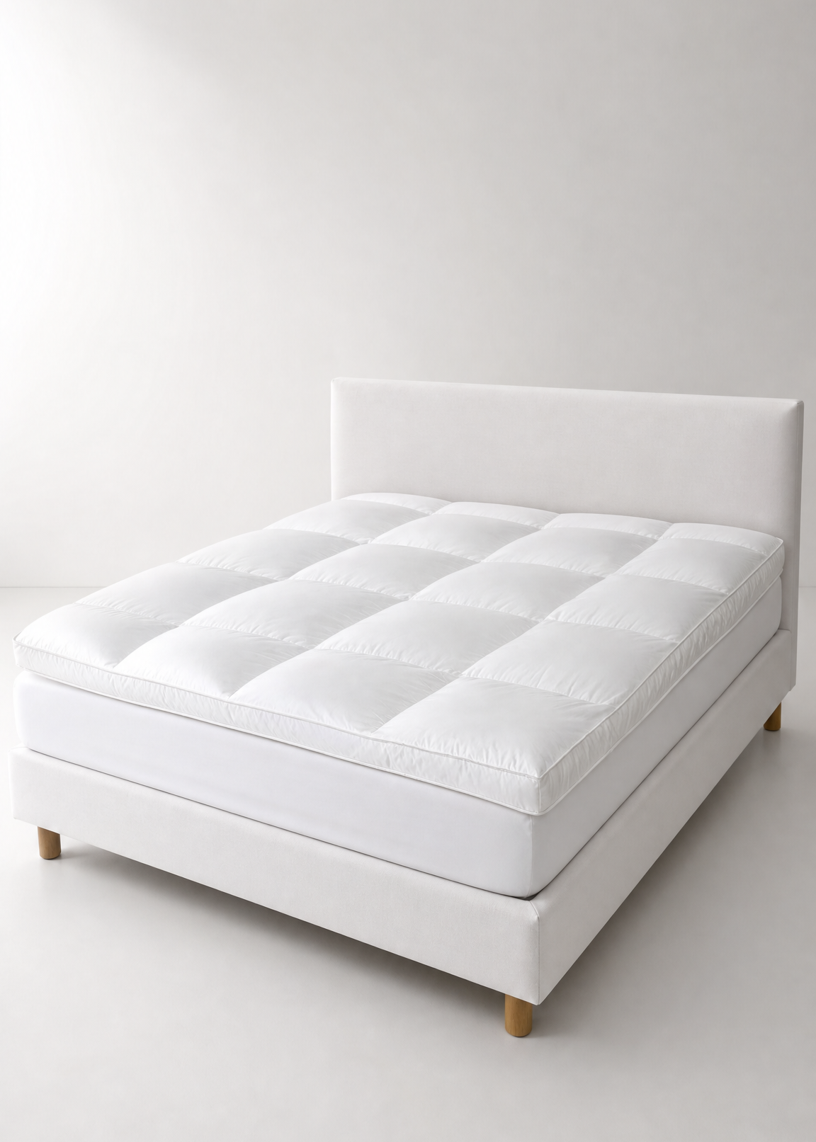 A white upholstered bed frame with a matching padded headboard and a quilted white mattress, photographed in a minimal white studio setting with light wood legs visible.