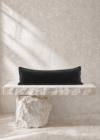 A black rectangular lumbar pillow displayed on a rustic stone bench against a textured beige wall with soft diagonal shadows.