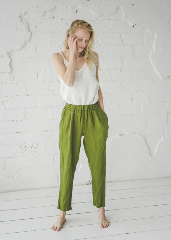 Woman wearing a white top and green pants against a white brick wall.