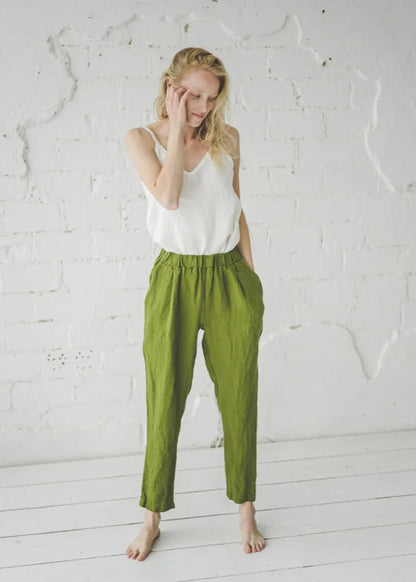 Woman wearing a white top and green pants against a white brick wall.
