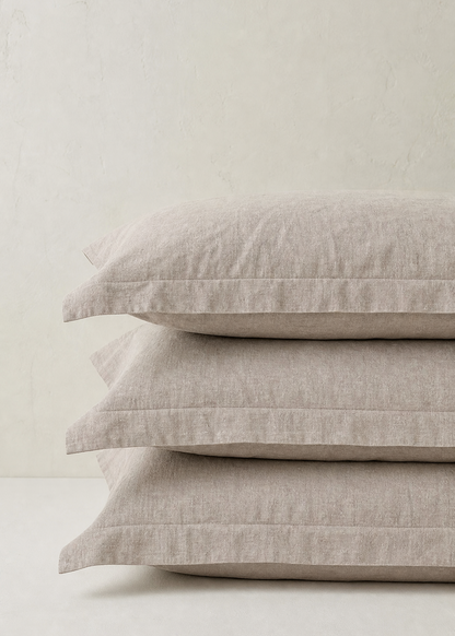 Three stacked linen pillows in a light oatmeal beige color against a neutral, textured off-white background.