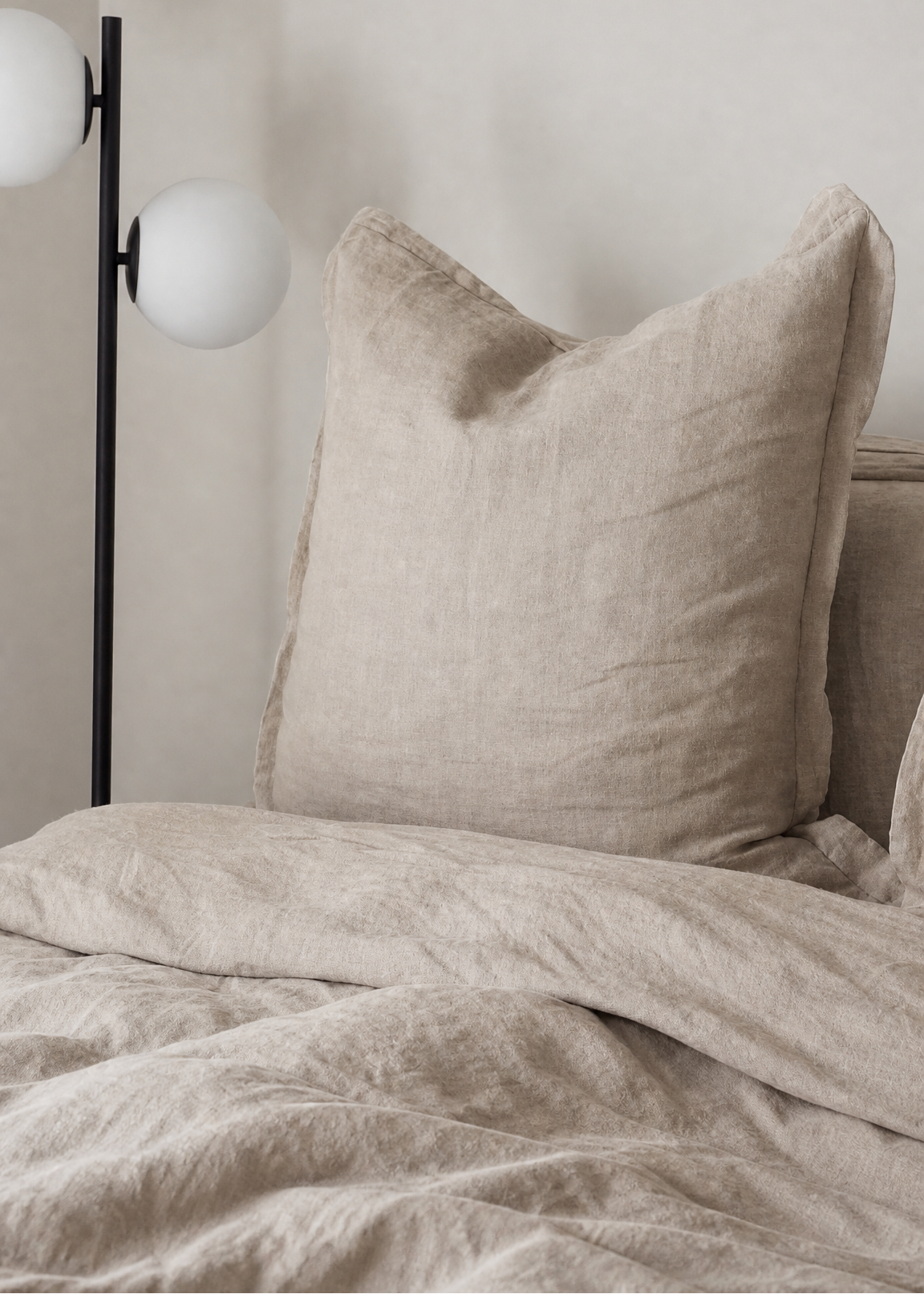 A close-up of a bed with textured beige linen bedding, featuring a large matching pillow and duvet, next to a modern black floor lamp with white globe shades against a neutral wall.