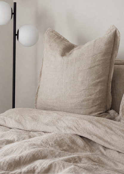 A close-up of a bed with textured beige linen bedding, featuring a large matching pillow and duvet, next to a modern black floor lamp with white globe shades against a neutral wall.