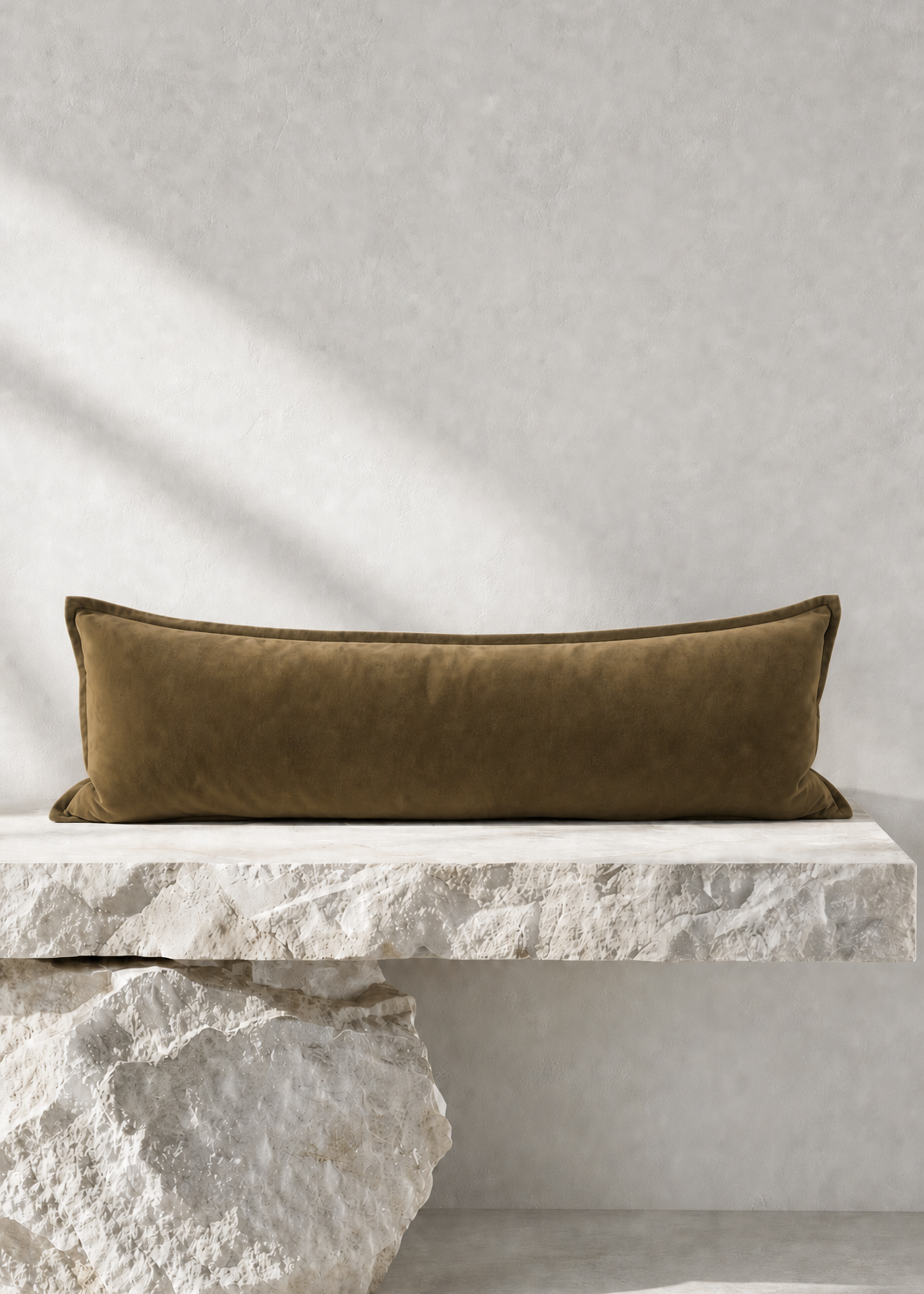 An olive green velvet rectangular lumbar pillow displayed on a white stone bench against a textured light gray wall with soft diagonal shadows.