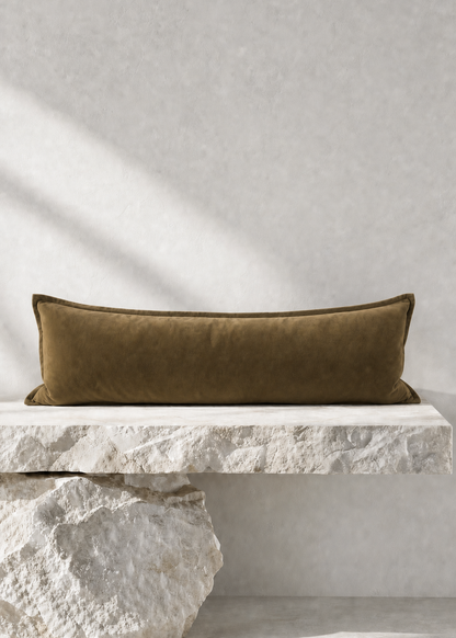 An olive green velvet rectangular lumbar pillow displayed on a white stone bench against a textured light gray wall with soft diagonal shadows.