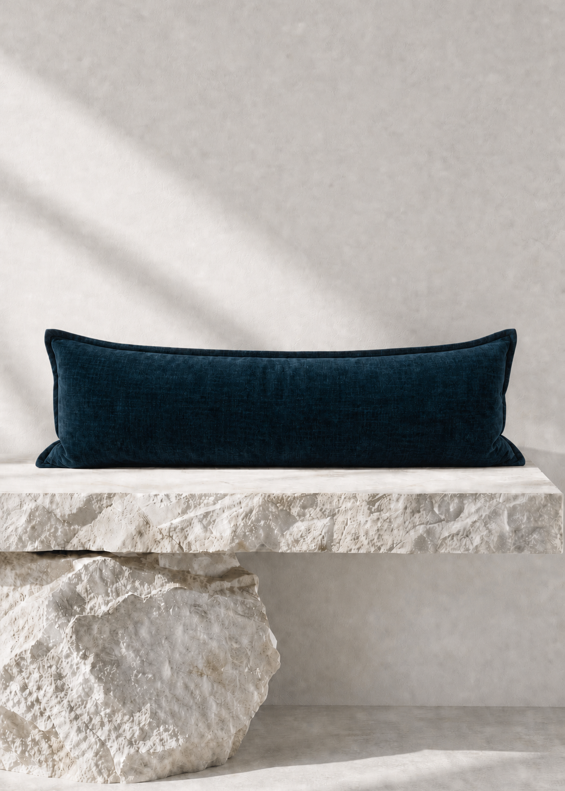 A dark navy blue velvet rectangular lumbar pillow displayed on a rustic white stone bench against a textured light gray wall with soft diagonal shadows.